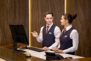 Two people in a hotel room at the front of a computer.